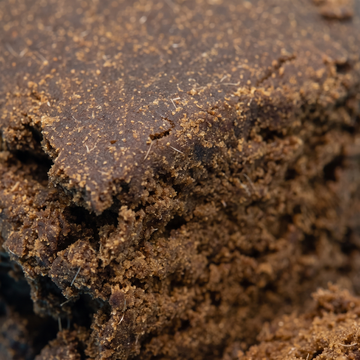 Makroaufnahme der Bruchkante des Aroma-Haschs: matte dunkelbraune Pressschicht über hellerem, sandbraunem und krümeligem Inneren, mit feinen hellen Pflanzenfasern